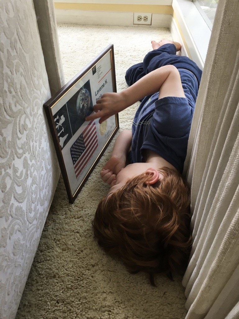Grace's little brother lying on the floor, studying a framed certificate with Mr. Peck's name and an American flag